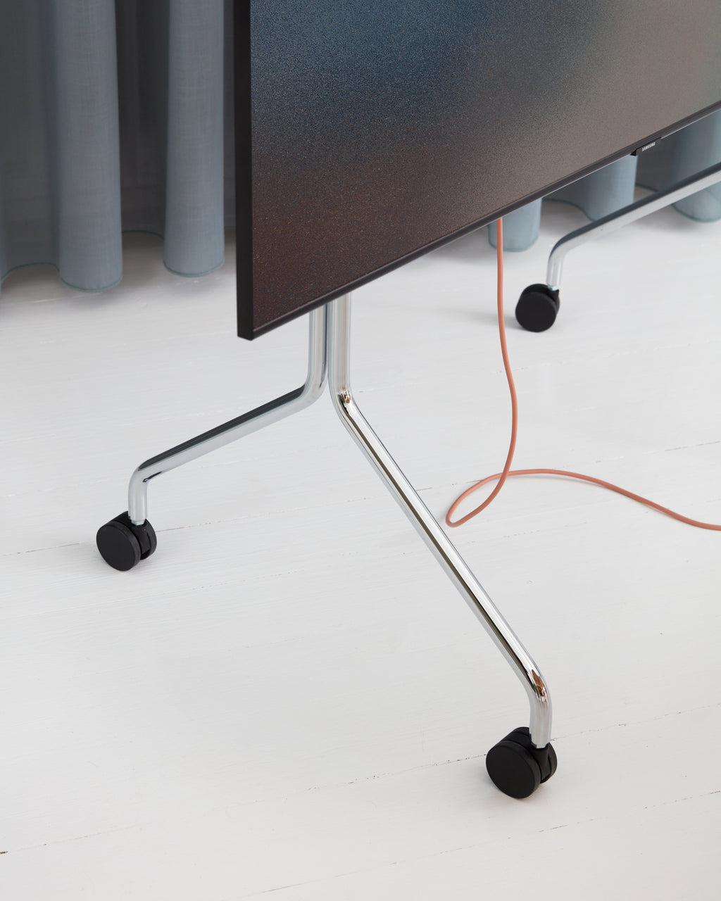Close-up of Moon in Chrome stand's angled legs and black locking wheels holding a TV on white studio flooring