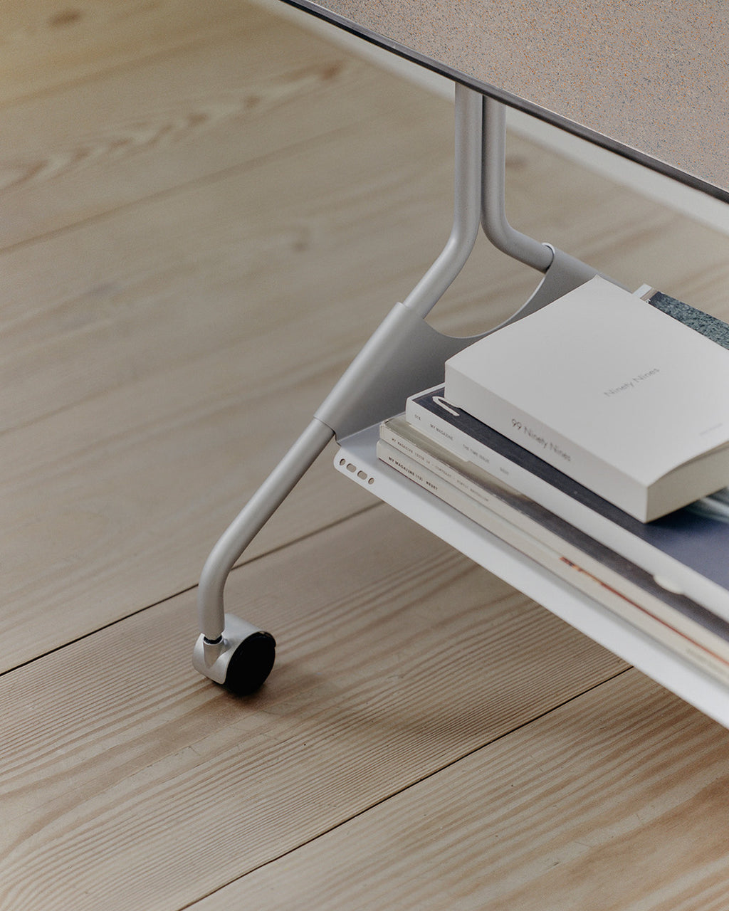 Close-up of Moon in Cloudy Silver stand's wheeled leg on pale wood floor, with books stacked on the lower shelf