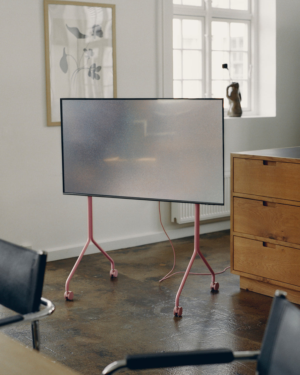 Moon Tall in Bubble Gum rolling TV stand holding a large screen in a bright office with oak drawers and leather chairs