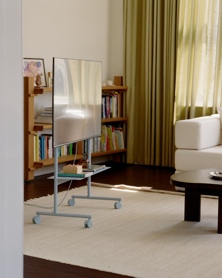 Rover stand in Powder Blue holding a TV on rolling casters in a sunlit living room with rug, bookshelves and sofa